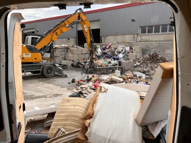Une excavatrice jaune déplace des débris devant un bâtiment industriel, vue depuis l'intérieur d'un véhicule rempli de meubles et de déchets.